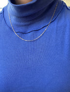 Someone wears a bright blue ribbed turtleneck top paired with the Adelene Paperclip Chain by LLUME Jewelry, a thin and delicate gold-plated necklace. The photo highlights the chest and neckline.