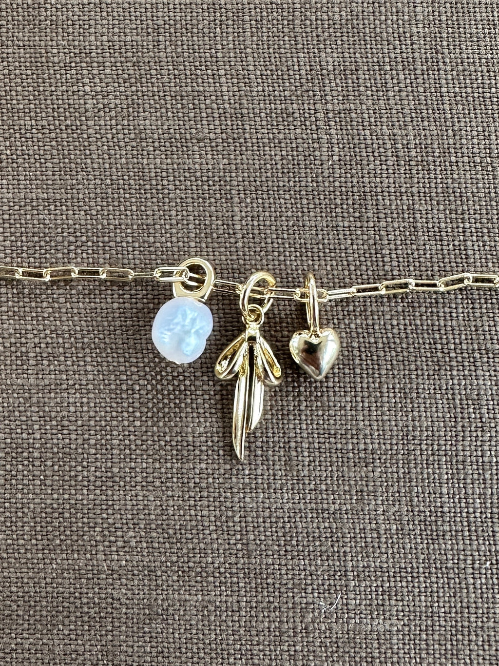 A gold chain bracelet with three gold-filled charms—a translucent flower, a wing, and a heart—from LLUME Jewelry rests on textured brown fabric, inspiring you to create your own Build a Charm Necklace or pair it with your favorite piece.