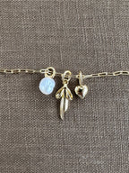 A gold chain bracelet with three gold-filled charms—a translucent flower, a wing, and a heart—from LLUME Jewelry rests on textured brown fabric, inspiring you to create your own Build a Charm Necklace or pair it with your favorite piece.