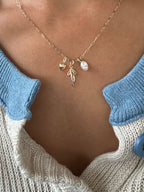 A close-up shows a person wearing LLUME Jewelry’s Build a Charm Necklace with gold filled heart, pearl, and leaf charms, styled over a cream sweater with blue collar and button for a personalized touch.