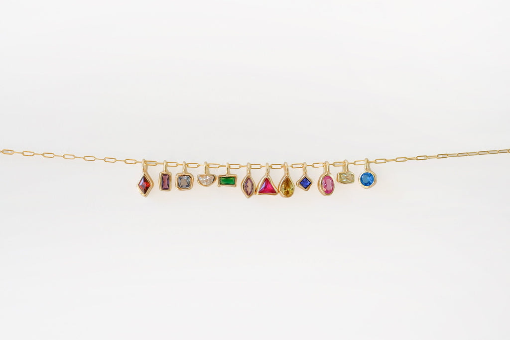 The Build a Birthstone Necklace by LLUME Jewelry features a gold chain with eleven unique, gold-plated gemstone charms in different shapes and settings, showcased against a plain white background.