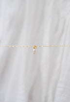 The Build a Charm Necklace by LLUME Jewelry features a delicate gold chain with a single small pearl pendant, shown on soft white fabric—ideal for layering or customizing with additional gold filled charms.