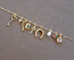 A gold chain by LLUME Jewelry showcases assorted gold filled charms—heart key, “mama” script, blue stone, and more—on a brown fabric backdrop, inspiring you to personalize your own Build a Charm Necklace.
