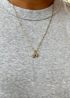 A person wears a light gray ribbed shirt and the LLUME Jewelry Build a Charm Necklace, featuring a gold chain with three gold-filled charms—one with red stones, one with a green stone, and one heart-shaped—for a personalized look.