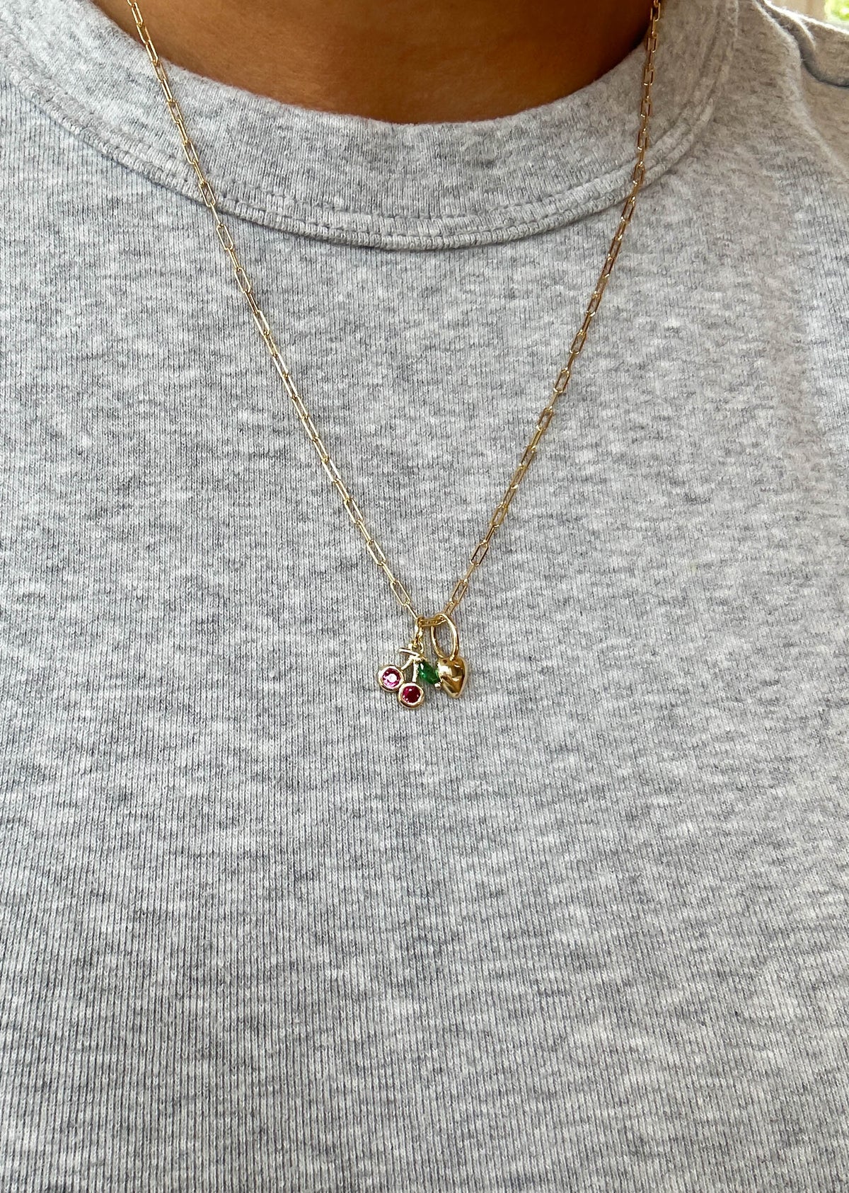 A person wears a light gray ribbed shirt and the LLUME Jewelry Build a Charm Necklace, featuring a gold chain with three gold-filled charms—one with red stones, one with a green stone, and one heart-shaped—for a personalized look.