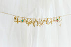 The Build a Charm Necklace by LLUME Jewelry features a gold chain adorned with gold filled charms—key, horseshoe, flower, heart, and birds—against a white fabric background.