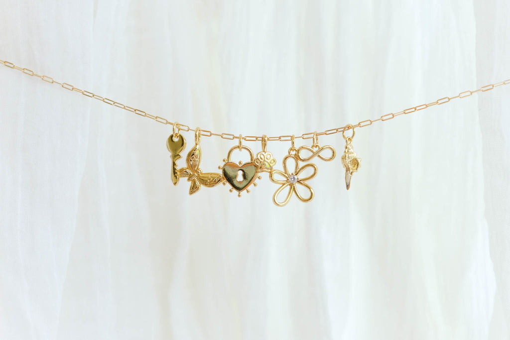 The Build a Charm Necklace by LLUME Jewelry features six unique gold filled charms—key, leaves, heart, paw print, gemmed flower, and ornate pendant—on a gold chain, elegantly displayed on soft white fabric for a truly personalized accessory.