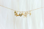 The Build a Charm Necklace by LLUME Jewelry features six unique gold filled charms—key, leaves, heart, paw print, gemmed flower, and ornate pendant—on a gold chain, elegantly displayed on soft white fabric for a truly personalized accessory.