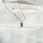 The Taylor Necklace by LLUME Jewelry, featuring a rectangular sapphire blue gemstone pendant with gold plating, is displayed on white textured fabric.
