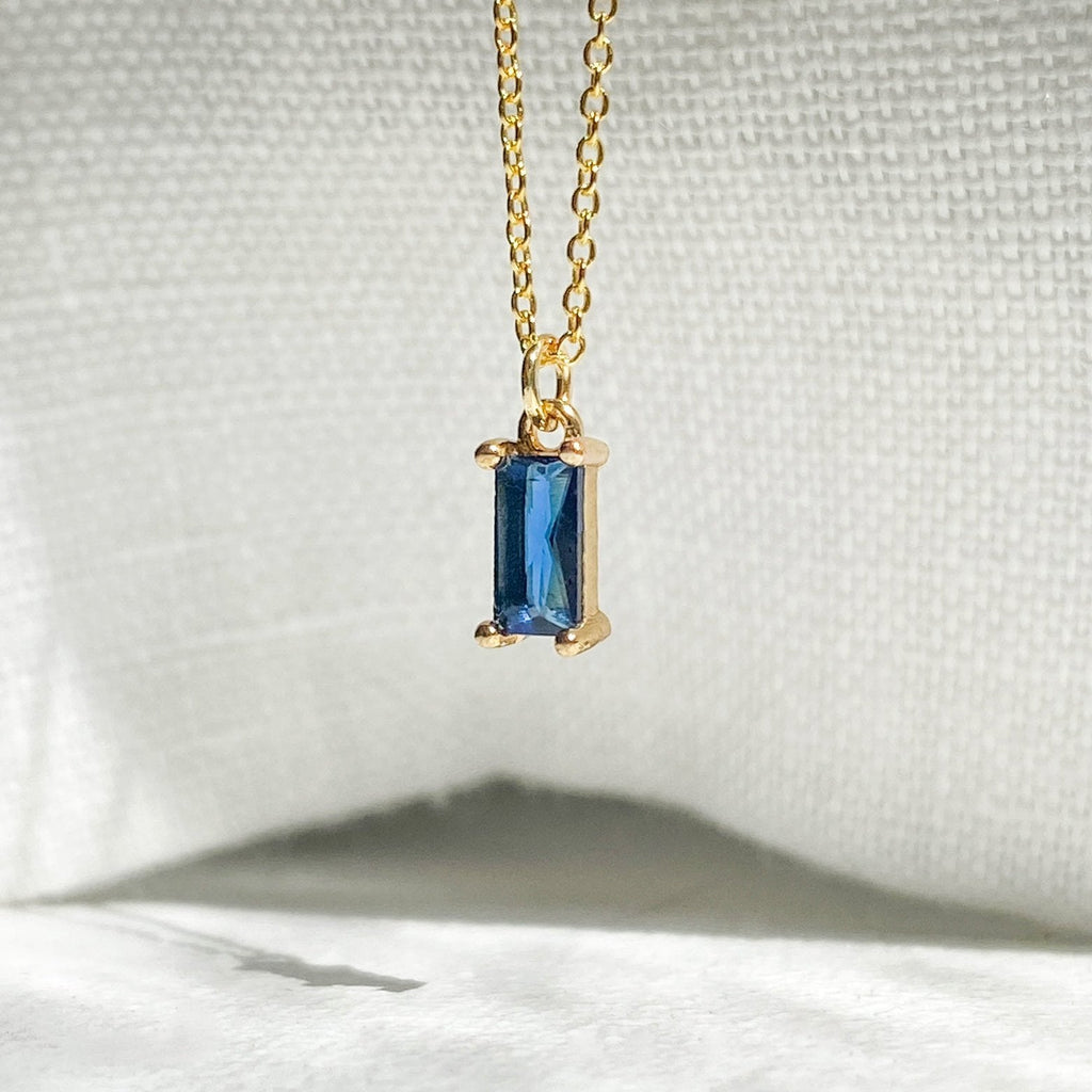 The Taylor Necklace by LLUME Jewelry features a gold plated chain with a rectangular sapphire blue gemstone pendant, shown on textured white fabric under soft lighting—a dainty charm perfect for any occasion.