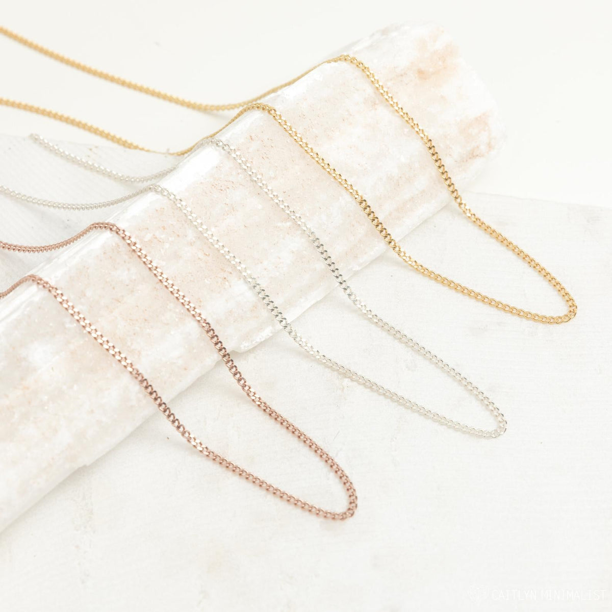 A variety of curb chain necklaces in gold, silver, and rose gold finishes displayed draped across a textured cream prop.