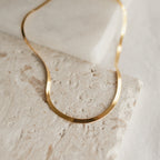 A close-up image of a thick herringbone necklace in yellow gold resting in gentle curves on a textured beige stone surface. The necklace displays a fluid, flat silhouette with fine, closely linked metalwork that gives it a smooth and reflective finish.