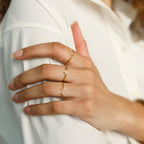 A hand displays three Modern Initial Birthstone Rings—customizable gold bands featuring small letters—worn against a white shirt.