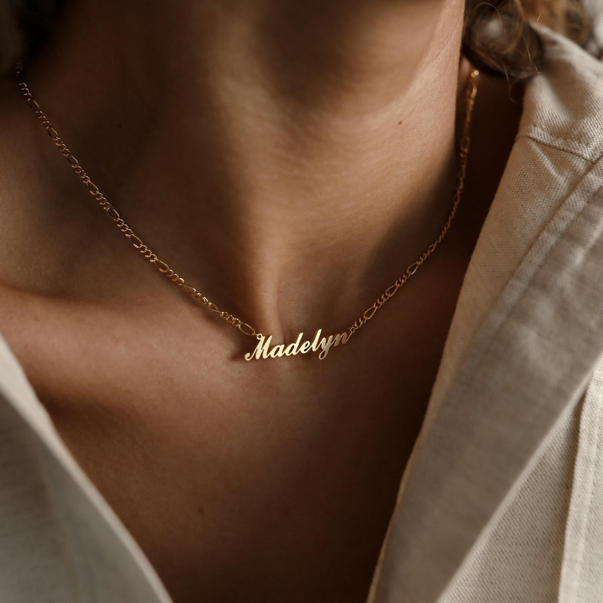 A minimal style gold Carrie Name Necklace in Figaro Chain featuring the name Madelyn, worn by someone in a cream-colored shirt.