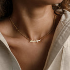 A woman wears the Carrie Name Necklace in Figaro Chain, featuring the name Madelyn in cursive gold letters.