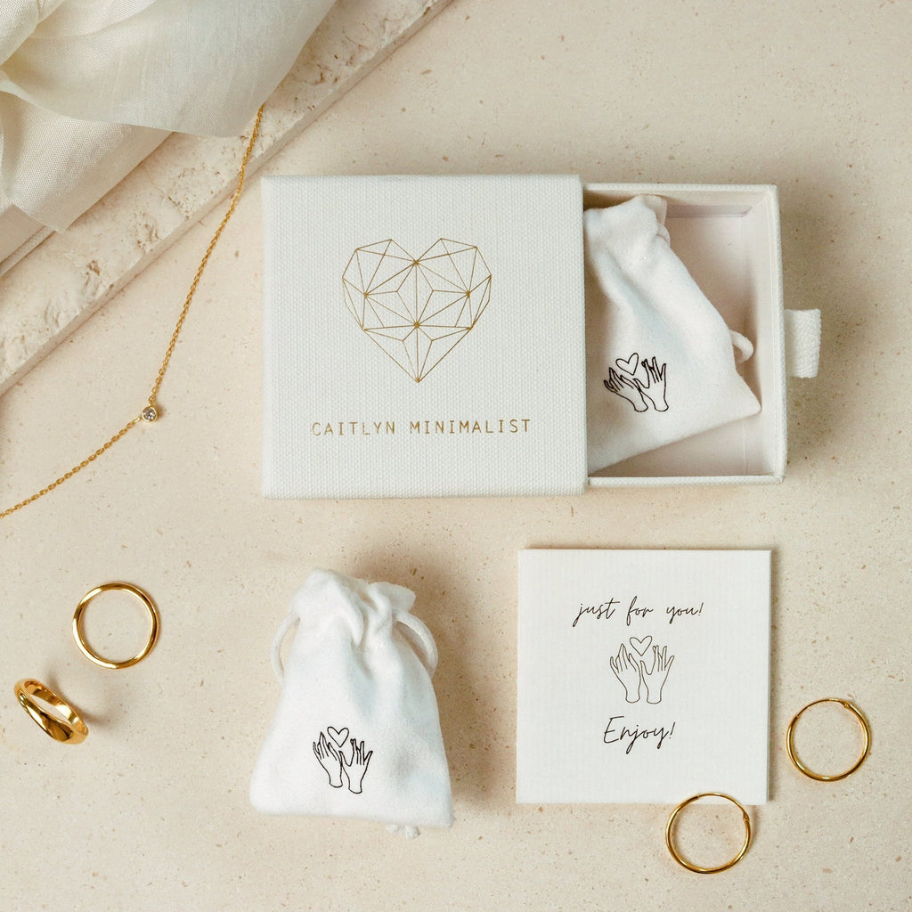 Mellow Name Heart Necklace displayed on a beige surface, featuring a dainty chain and packaged in a box labeled Caitlyn Minimalist.