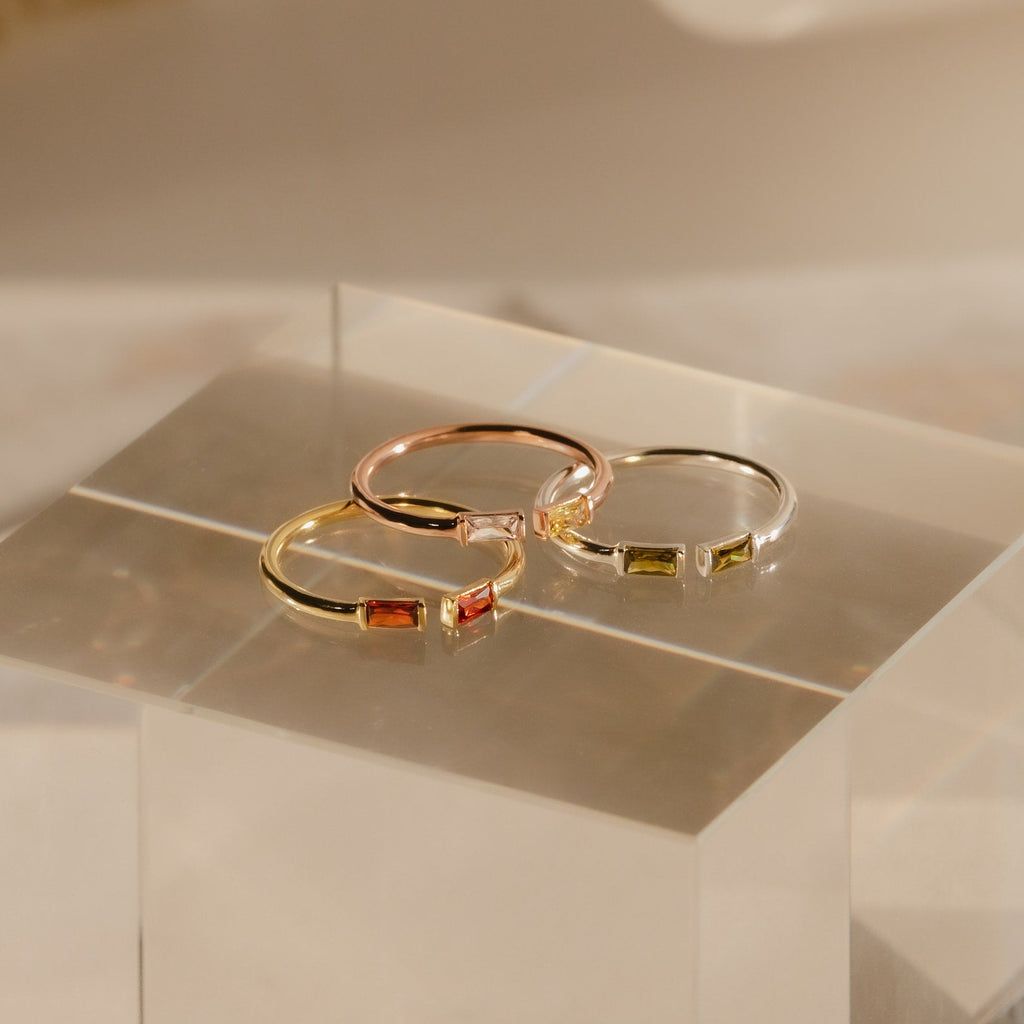 The Duo Baguette Birthstone Ring, a minimalist piece with colored rectangular stones, is displayed on a clear glass cube under soft lighting—a perfect personalized gift with customized gemstones.