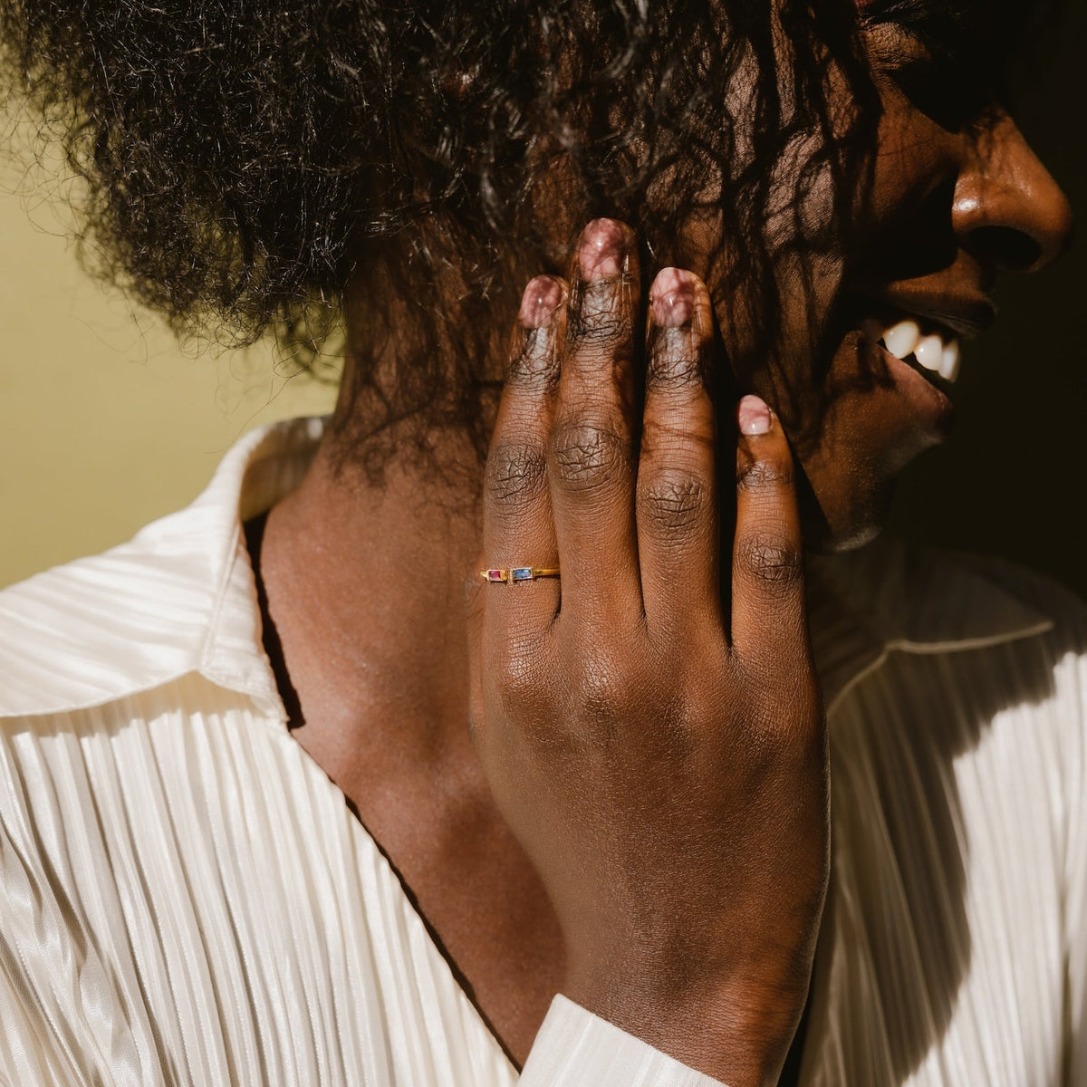 A smiling person touches their cheek, wearing the Duo Baguette Birthstone Ring with personalized gemstones, paired with a light blouse.