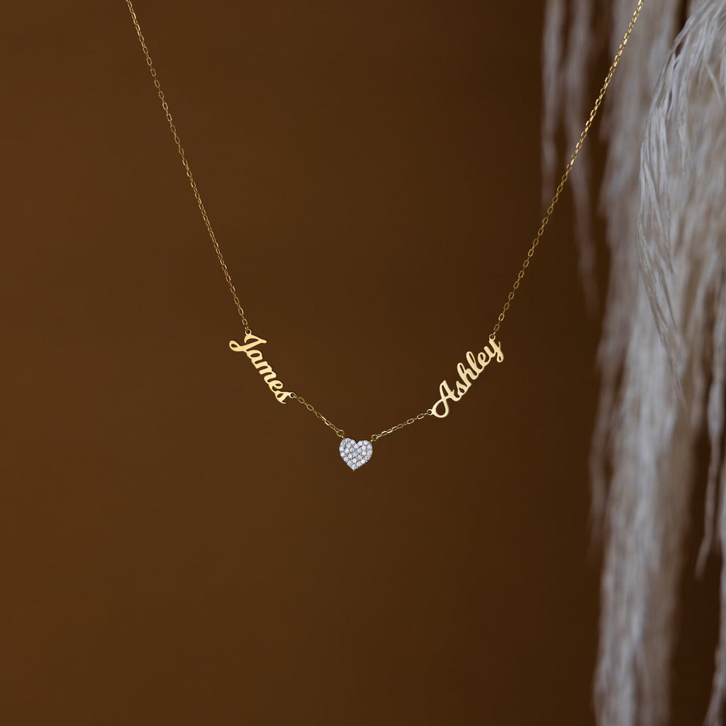 The Mellow Name Heart Necklace features "James" and "Ashley" connected by a small heart charm on a delicate gold chain, displayed against a brown background.