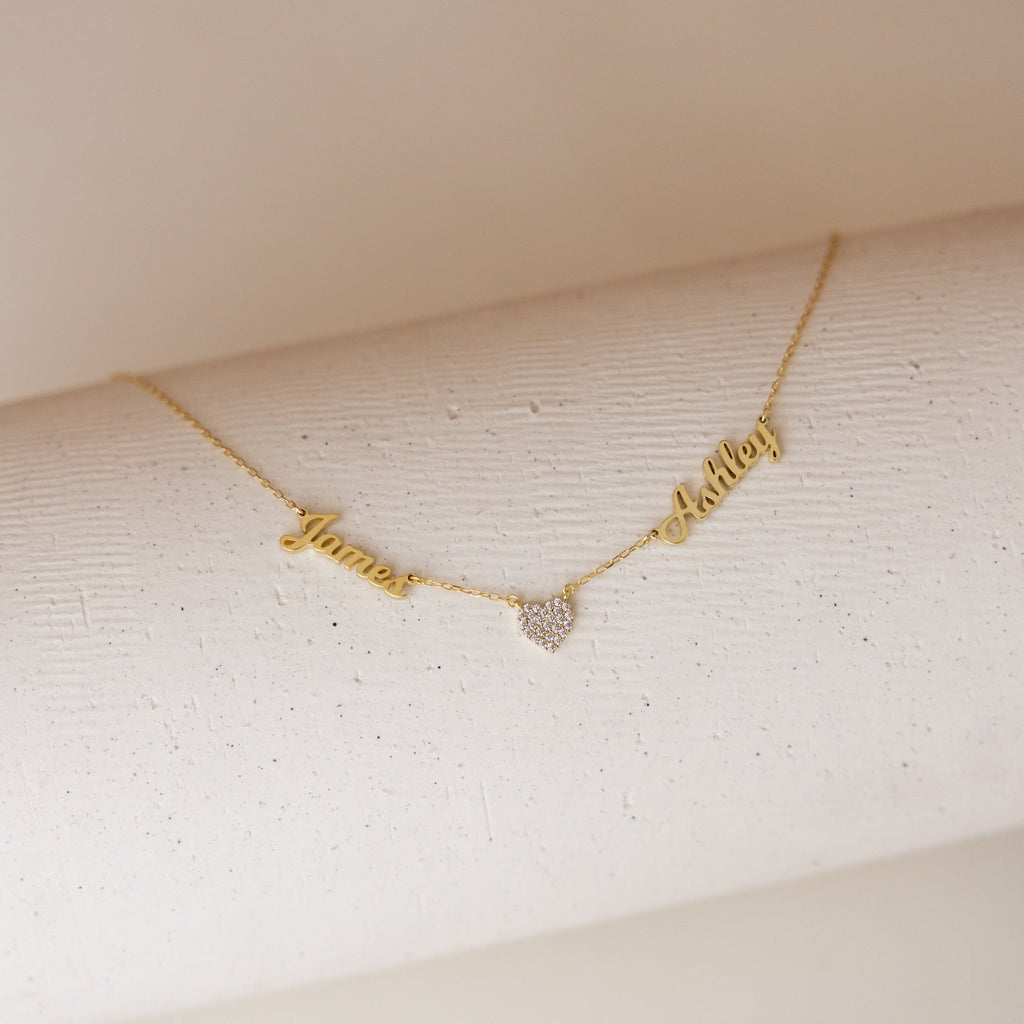 The Mellow Name Heart Necklace features gold personalized names, James and Ashley, connected by a small heart pendant on a dainty chain, displayed on a white textured surface. Perfect for adding a sweet touch to any outfit.