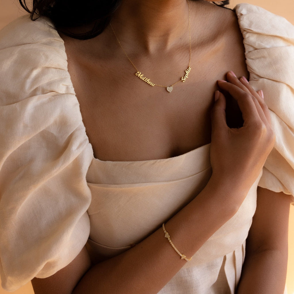 Woman in a cream puff-sleeve dress wearing a gold bracelet and the Mellow Name Heart Necklace, a dainty personalized piece featuring two charms and a heart.