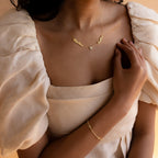 Woman in a cream puff-sleeve dress wearing a gold bracelet and the Mellow Name Heart Necklace, a dainty personalized piece featuring two charms and a heart.