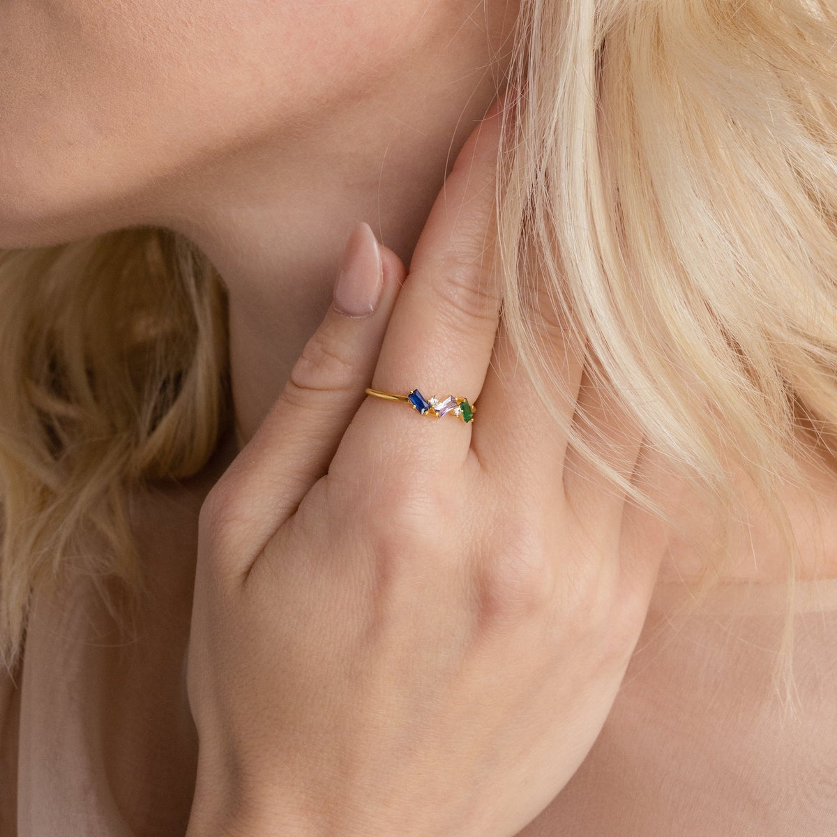 A woman with blonde hair touches her neck, wearing the Cluster Baguette Birthstone Ring adorned with colorful gemstones on her finger.