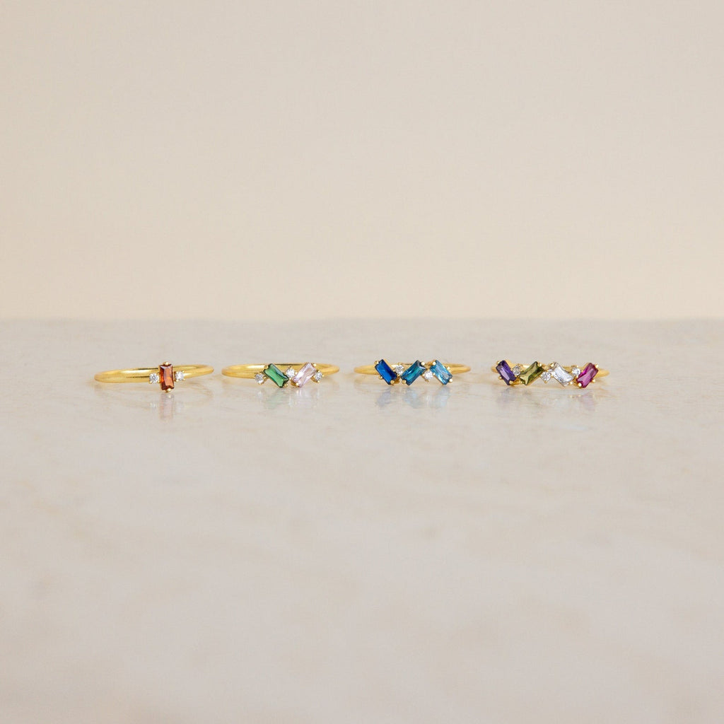 Five Cluster Baguette Birthstone Rings with colorful gemstones are arranged in a row on a light marble surface against a beige background, perfect as birthstone or personalized statement rings.