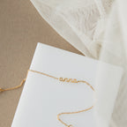 The Petite Tiny Name Bracelet showcases "anna" on a delicate chain, set against a white surface with beige fabric and mesh—ideal for minimal jewelry enthusiasts.