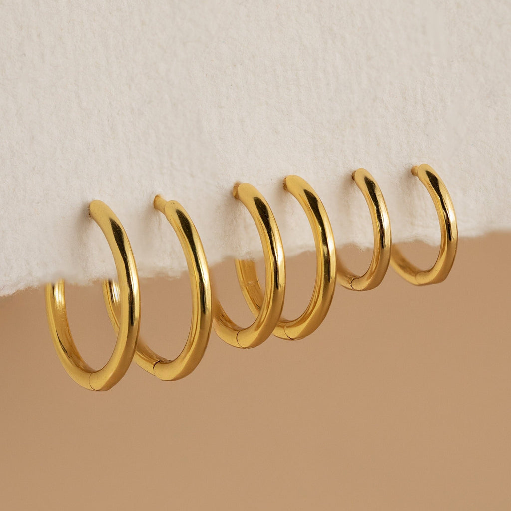 Six gold Mini Bold Hoops from our collection hang on the edge of a white textured surface, showcasing their varying sizes and timeless style.
