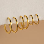 Six gold Mini Bold Hoops from our collection hang on the edge of a white textured surface, showcasing their varying sizes and timeless style.