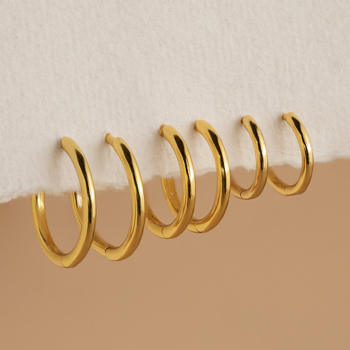 Six gold Mini Bold Hoops from our collection hang on the edge of a white textured surface, showcasing their varying sizes and timeless style.