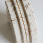 The Marie Chain by LLUME Jewelry, an 18k gold-filled necklace, is displayed draped over a polished marble surface with white, beige, and brown veining and carved grooves.