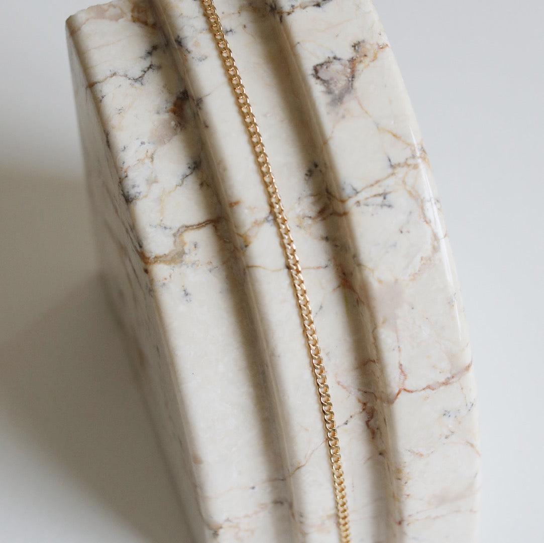 The Marie Chain by LLUME Jewelry, an 18k gold-filled necklace, is displayed draped over a polished marble surface with white, beige, and brown veining and carved grooves.