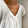 A person wears a white, pleated V-neck blouse and LLUME Jewelry’s Celia Necklace; soft light enhances the fabric’s texture and the wearer’s collarbones.