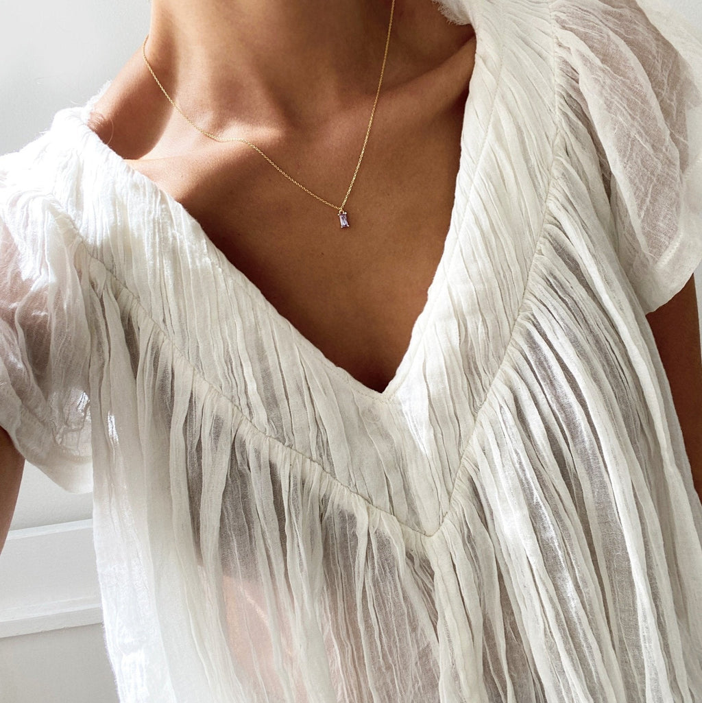 A person wears a sheer white pleated blouse with a deep V-neckline and the Celia Necklace by LLUME Jewelry in gold vermeil. The close-up image highlights the necklace’s subtle shine on the upper chest and neck.