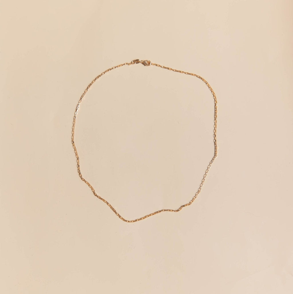 The Harlow Chain by LLUME Jewelry, a delicate 18k gold filled necklace with a subtle twisted design, is arranged in an uneven oval on a light beige background.