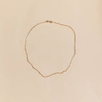 The Harlow Chain by LLUME Jewelry, a delicate 18k gold filled necklace with a subtle twisted design, is arranged in an uneven oval on a light beige background.