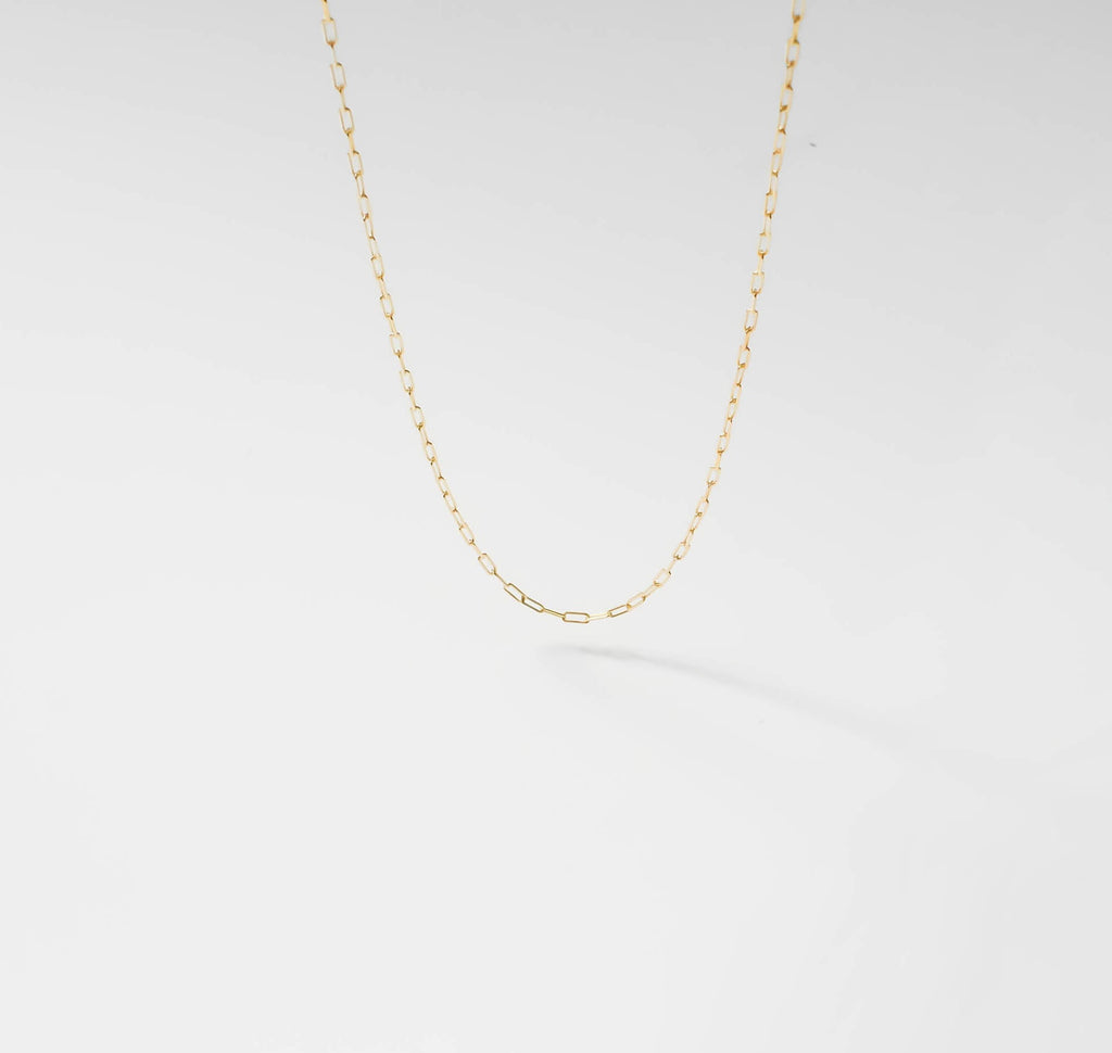 The Adelene Paperclip Chain by LLUME Jewelry features small, oval gold-plated links. Shown against a white background, the adjustable necklace forms a gentle curve and casts a subtle shadow below.