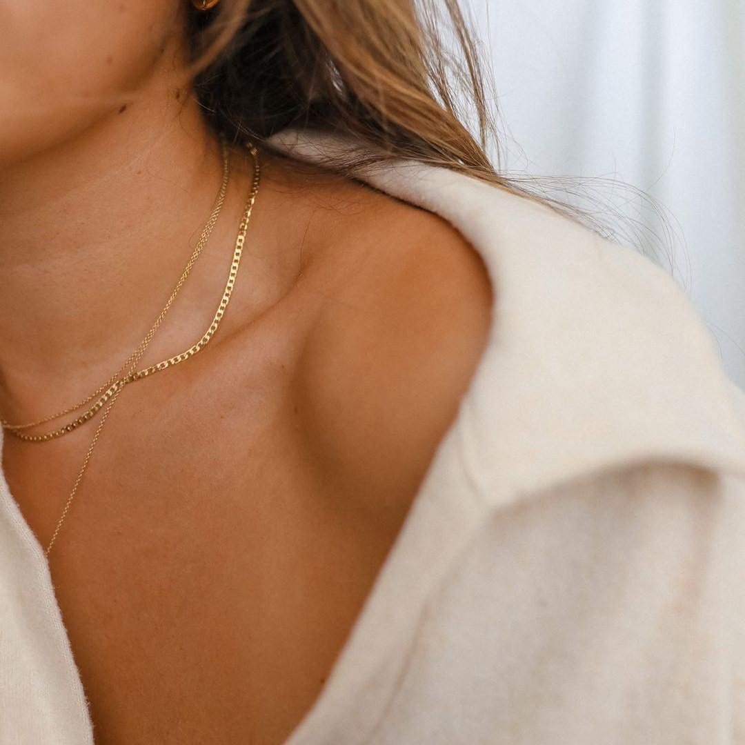 Close-up of a woman in a cream off-shoulder top wearing LLUME Jewelry's waterproof Marie Chain, an 18k gold-filled necklace. Only part of her face, neck, shoulder, and hair are visible against a softly blurred background.