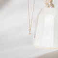 The Daylight Necklace by LLUME Jewelry, featuring a delicate gold-plated chain and a small rectangular clear gemstone pendant, is displayed in front of a white ceramic vase against a soft, minimal background.