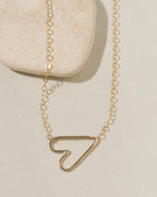 Delicate gold chain necklace with a sideways open-outline gold heart pendant on cream surface with stone pebble