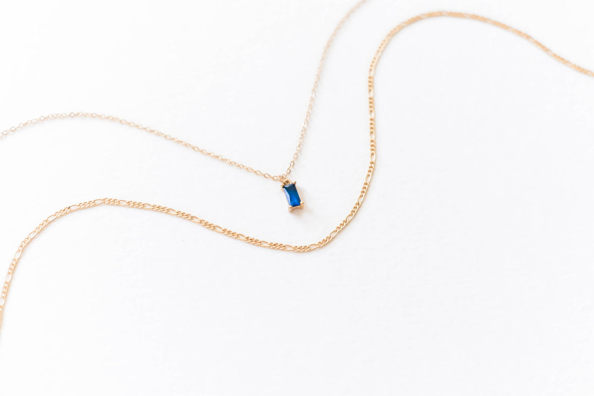 The LLUME Jewelry Taylor Daily Set features a delicate gold pendant necklace with a thin chain and small rectangular blue gemstone, paired with a second slim gold chain—ideal for an elegant layered look.